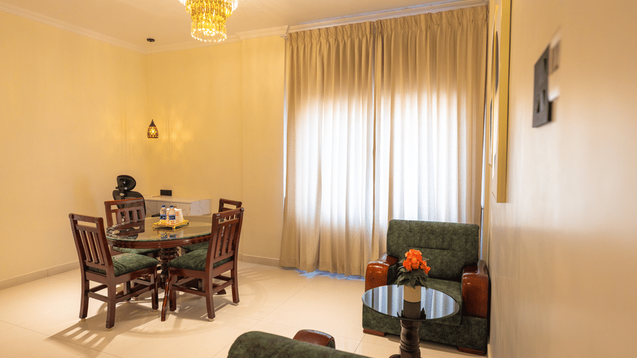 Suite living area at DSF Grand Plaza, Tuticorin with a round dining table, 4 wood chairs, a crystal chandelier, green armchairs, and floor-length curtains.