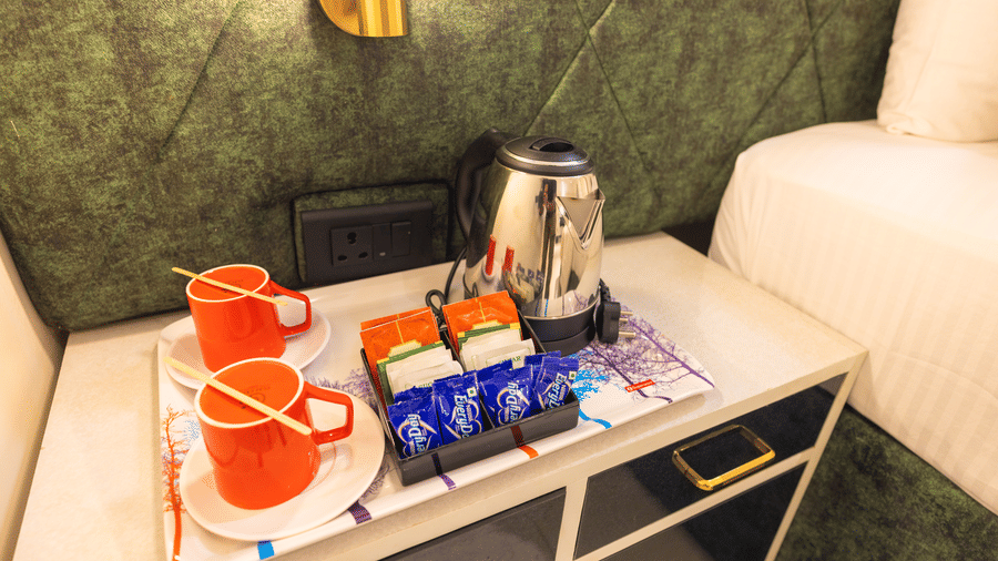 Tea and coffee station at DSF Grand Plaza, Tuticorin with a kettle, 2 red mugs on saucers, and sachets of coffee, tea, and sugar on a bedside tray.