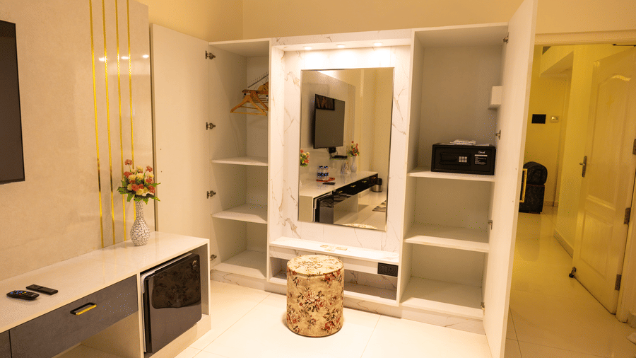Wardrobe and dressing area at DSF Grand Plaza, Tuticorin with open shelves, hanging space, an in-room safe, a mirrored vanity, and a floral ottoman.
