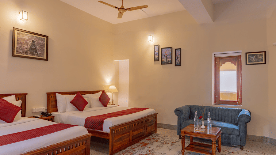 A spacious room at Estherea Bagh, Ranthambore, containing 2 wooden beds with red accents, an upholstered sofa, and a ceiling fan.
