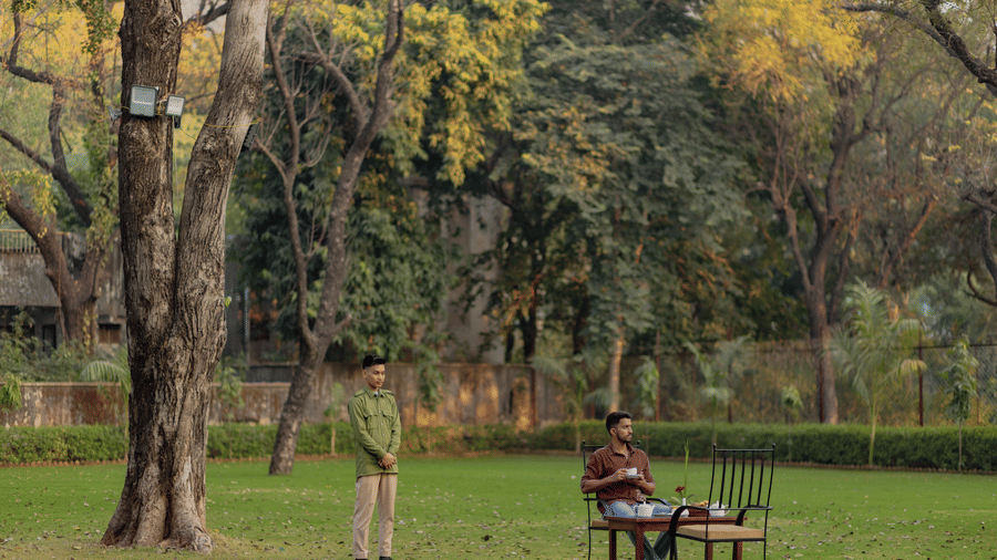 A person sitting at a tea table on the vast green lawn of Estherea Bagh, Ranthambore, shaded by tall trees.