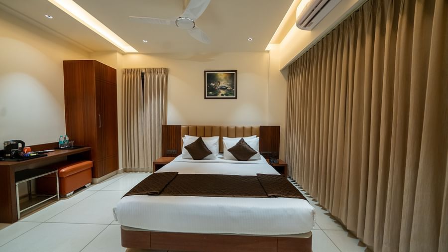 A front view of the bed at Hari Govinda's Express, Vijayanagar, with a photo frame above the headboard. To the left, a wooden cupboard stands beside a table with essentials, while on the right, an AC is mounted above the fully drawn curtains.
