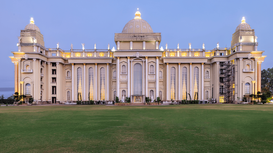 Vast, ornate, pale-coloured building with a central dome and grand architecture, illuminated against a twilight sky, set on a lawn at Hotel Hukam's Lalit Mahal.