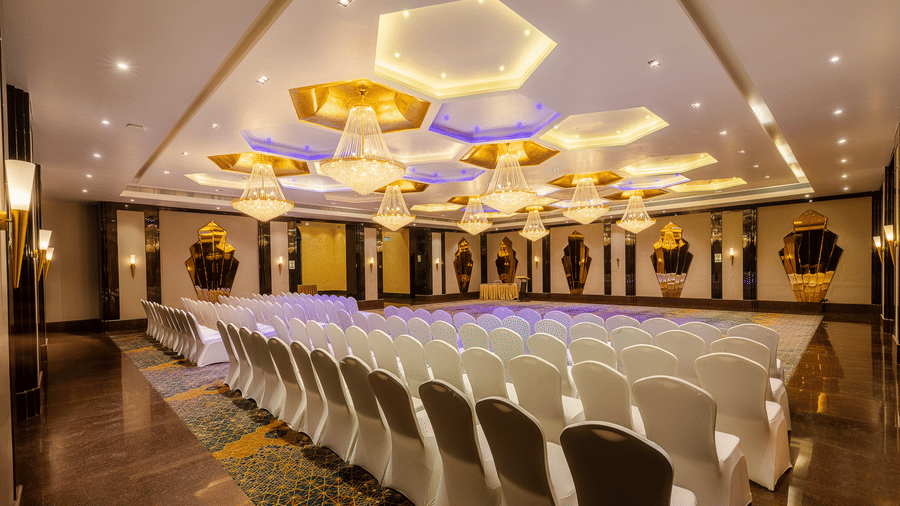 Banquet hall set with rows of white chairs facing a stage area, brightly lit by impressive hexagonal ceiling fixtures at Hotel Hukam's Lalit Mahal.