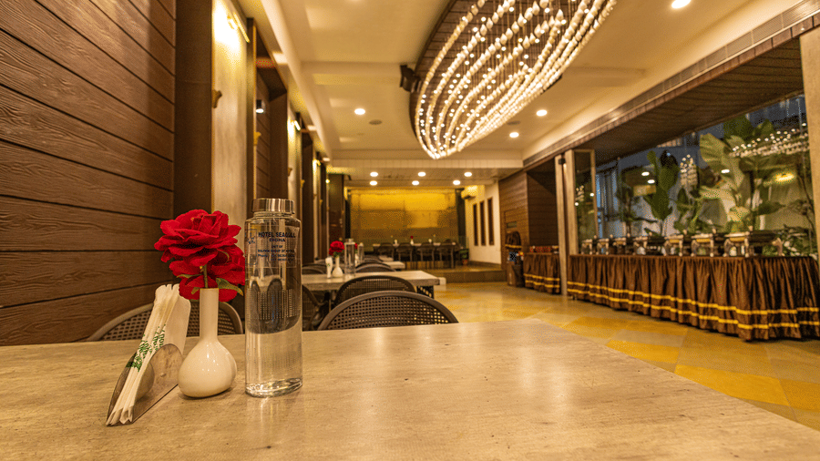 A dining area at Hotel Seagull Digha with a table, a flower in a vase, chairs, ceiling lights, and a decorative chandelier overhead.