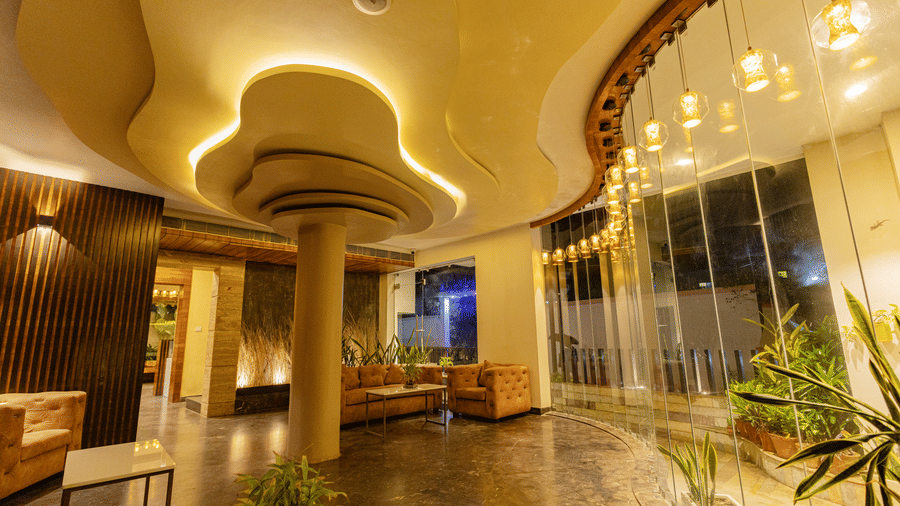 A lobby area with 2 sofas, a table, ceiling lights, plants, and glass walls at Hotel Seagull Digha.