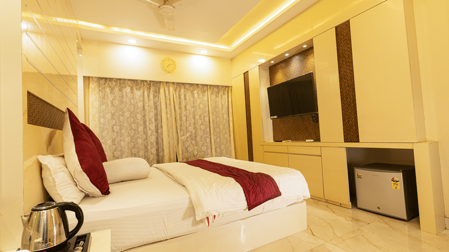 A bedroom with a king size bed, a bedside table with an electric kettle, a television, a mini fridge, and curtains at Hotel Seagull Digha.