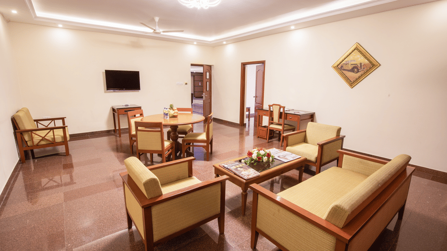 A spacious living area within a hotel suite featuring wooden armchairs a coffee table and a dining set - Jenneys Residency, Coimbatore