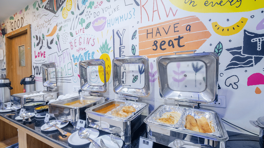 Metal food warmers with open lids line a counter next to a wall covered in drawings and text at Lemonridge by Monday Hotels - Kukatpally's restaurant Curry Leaf.