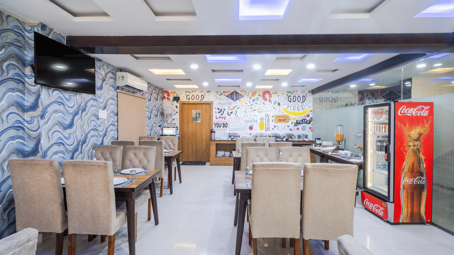 A view inside the restaurant at Lemonridge by Monday Hotels - Kukatpally featuring rows of tables, high back chairs, and a red beverage refrigerator.