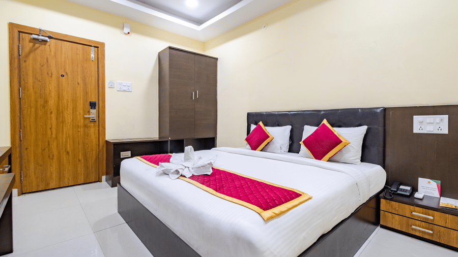 A room at Lemonridge by Monday Hotels - Kukatpally features a large bed, a tall wardrobe, a wooden entrance door, and bedside tables.