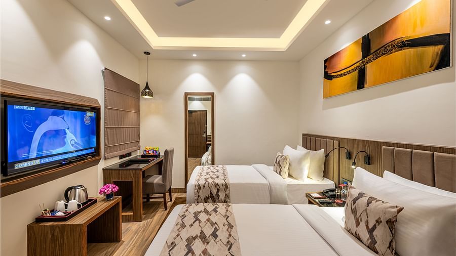 A hotel room with two twin beds, a flat-screen TV, and a work desk. It features a unique tray ceiling with lighting, a ceiling fan, and colourful abstract artwork - Lords Eco Inn Dehradun. 