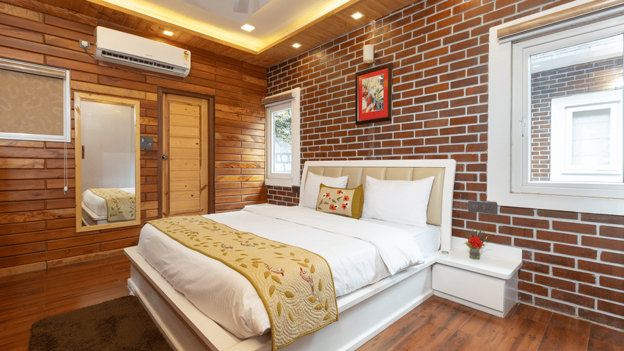 A well-lit bedroom showcasing the red brick and wooden wall behind the large, comfortable bed at Megha Resort & Villas, Dehradun.