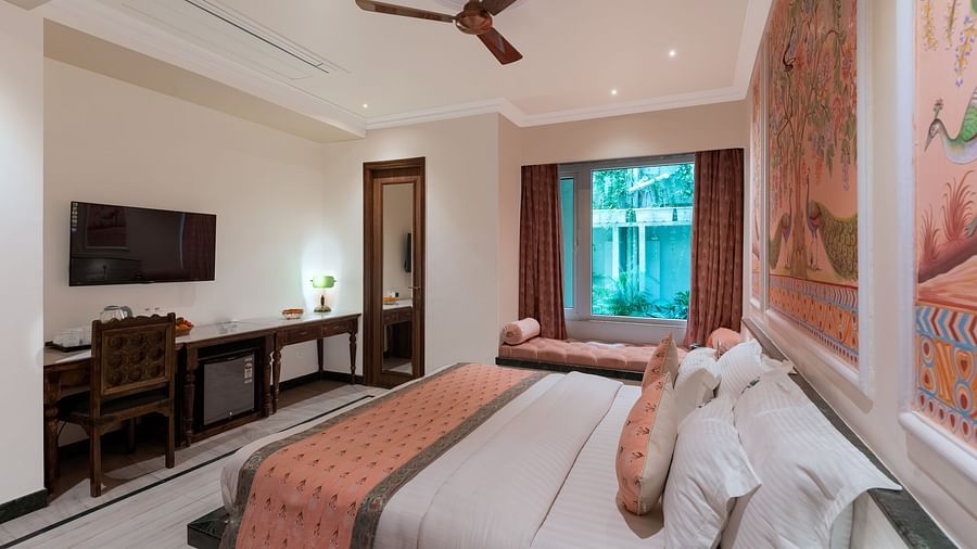 A headboard view of a spacious bedroom at Panna Vilas Palace, displaying a wall-mounted television with a wooden table underneath and large windows inviting natural light