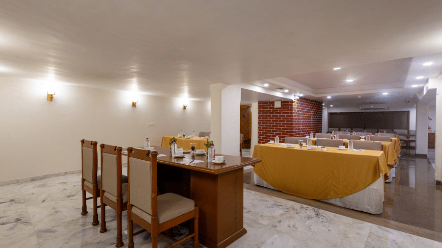 A well-lit hotel dining space showing a corner with wooden chairs, a table set for dining, and a yellow sofa at Pravasa Stay, Vadodara.