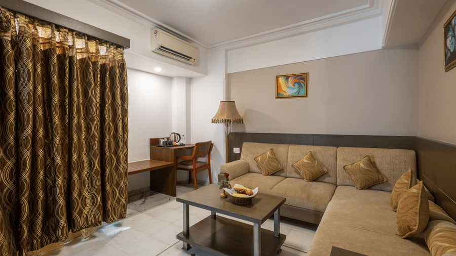 A hotel suite lounge with a large, beige L-shaped sofa, coffee table, and heavy, patterned brown curtains at Pravasa Stay, Vadodara.
