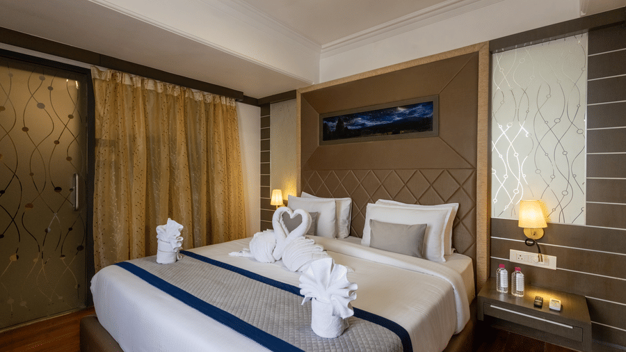 A hotel bedroom with a neatly made bed featuring towel swans and blue accents, wood panelling, and soft bedside lighting at Pravasa Stay, Vadodara.