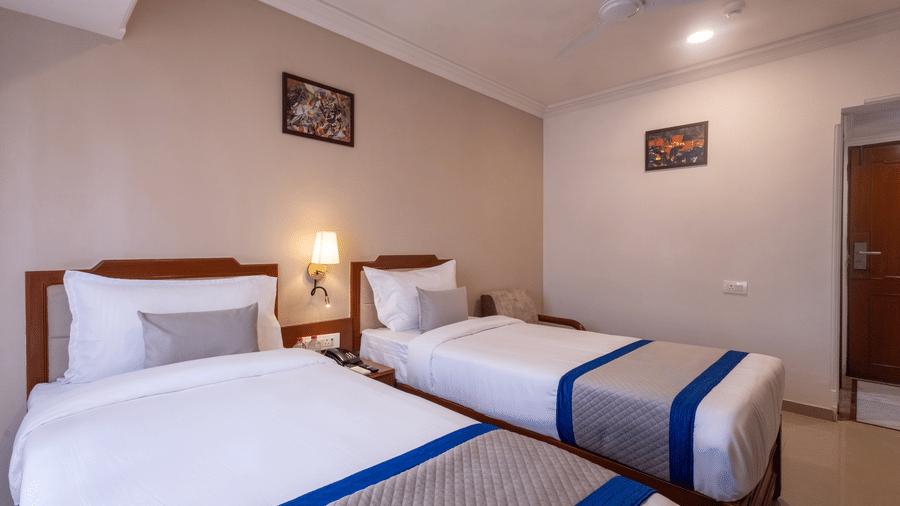 A bright hotel room with two single beds, each made up with white linen and a striking blue and grey throw at Pravasa Stay, Vadodara.
