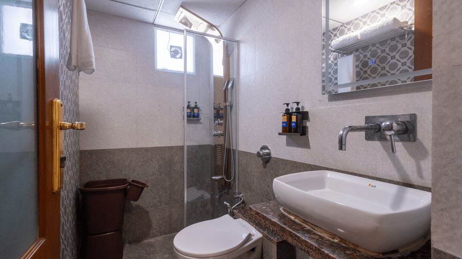 Modern hotel bathroom with a glass shower screen, wall-mounted basin, and warm lighting on tiled walls at Pravasa Stay, Vadodara.