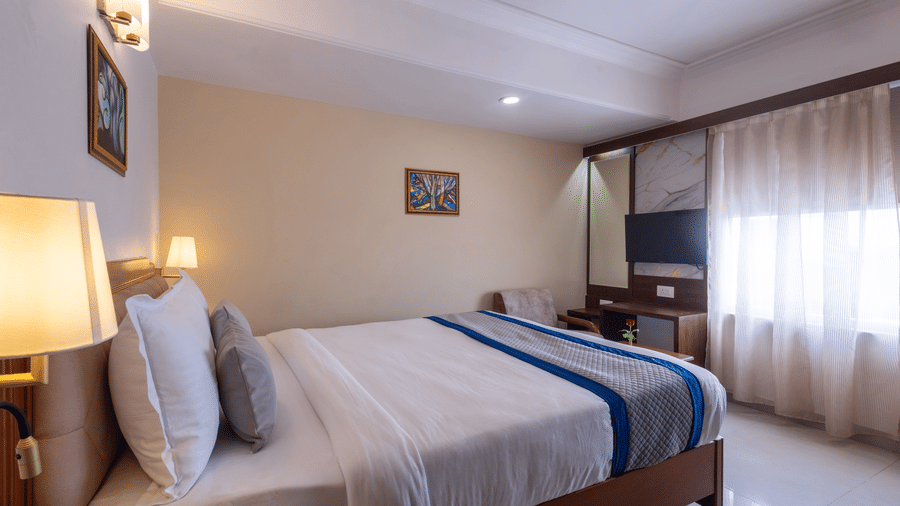 A warm, contemporary hotel room with a large bed and bright window, featuring wood accents and soft lighting at Pravasa Stay, Vadodara.