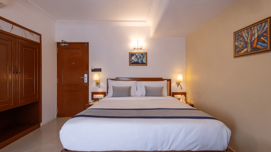 A view of the king-sized bed and headboard from the doorway, showing the wooden floor and warm bedroom lighting at Pravasa Stay, Vadodara.