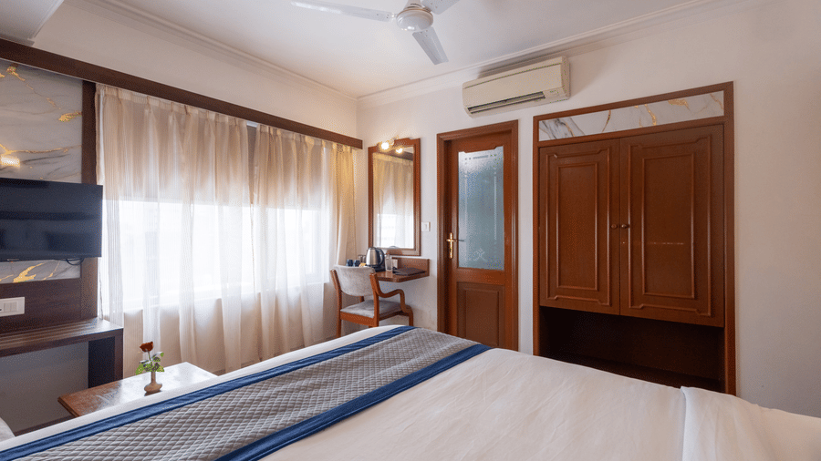 View across the bed towards a large wooden wardrobe and mirrored door, showing the room's spacious layout at Pravasa Stay, Vadodara.