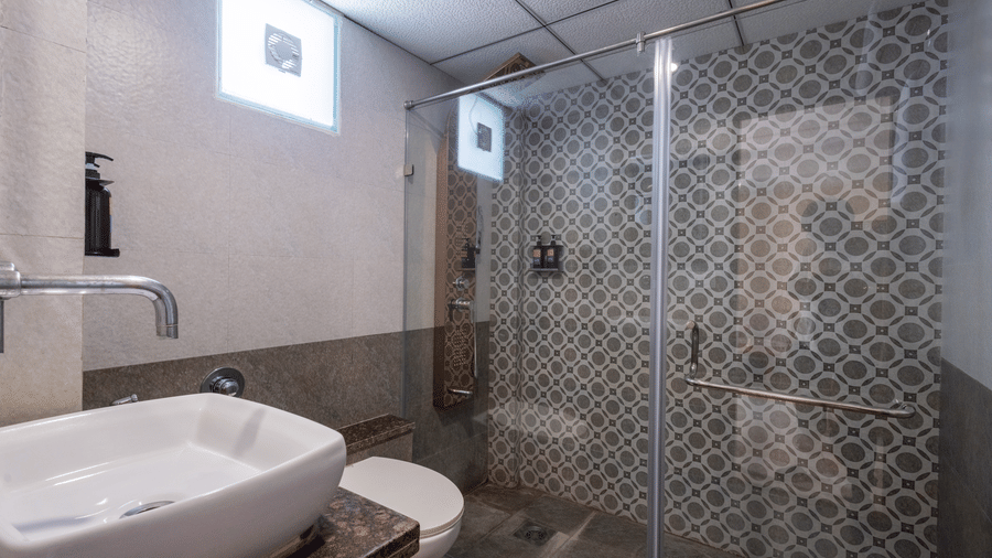 A sleek hotel bathroom shower area with a glass partition and decorative patterned grey and white wall tiles at Pravasa Stay, Vadodara.
