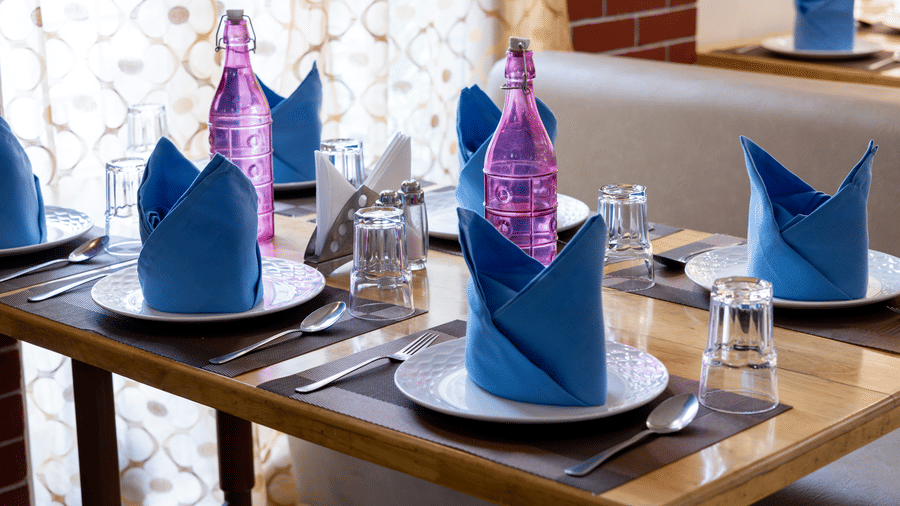 Close-up of a wooden table setting with white plates and striking blue and pink folded napkins in a well-lit dining area at Pravasa Stay, Vadodara.