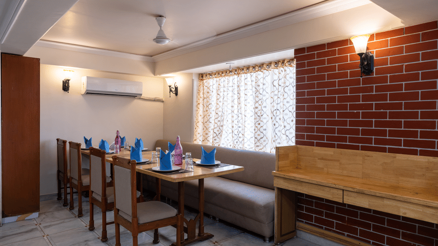 Restaurant seating area featuring wooden chairs, a long booth, exposed brickwork, and an air conditioning unit at Pravasa Stay, Vadodara.