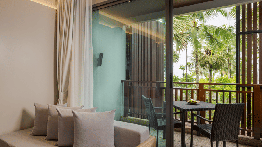 A balcony of Deluxe Jet Tub room with outdoor chairs and a small table, connected to an indoor seating area and surrounded by greenery at Ramada Palace.