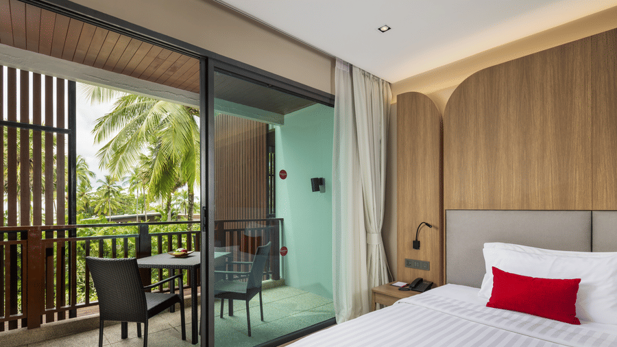 A view of a Jet Tub twin bedroom with a bed positioned beside sliding glass doors opening to a balcony with outdoor seating and greenery visible at Ramada Palace.