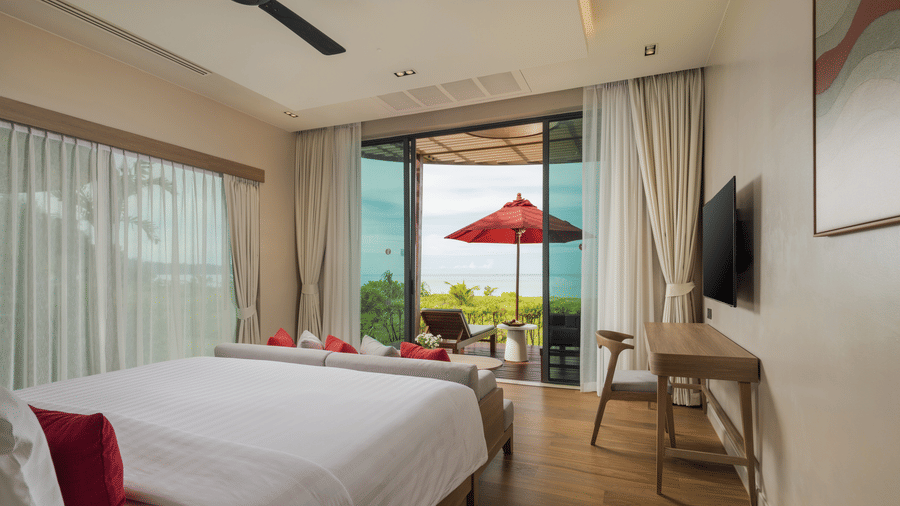 A bedroom of Oceanfront Villa with a large bed, writing desk, wall-mounted television, and balcony doors opening to outdoor seating at Ramada Resort.