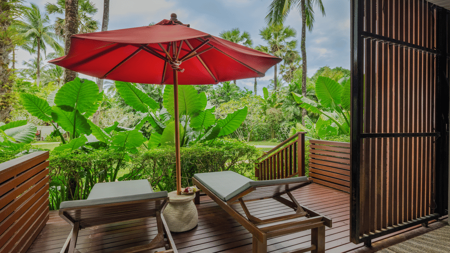 An outdoor balcony of Jet Tub Lanai room with wooden loungers, a small table, and a red umbrella surrounded by plants at at Ramada Resort.