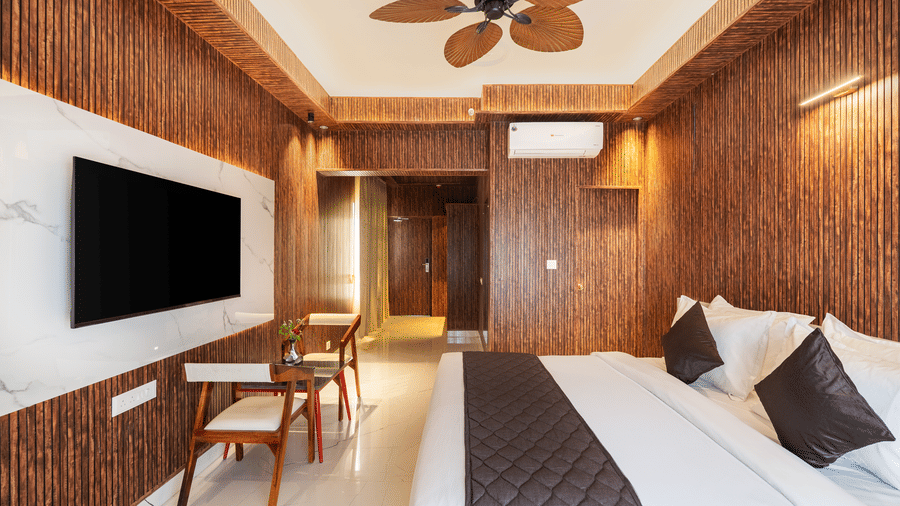 Hotel bedroom interior with a king-size bed, dark wooden wall panelling, a dark floor runner, and a large wall-mounted TV.