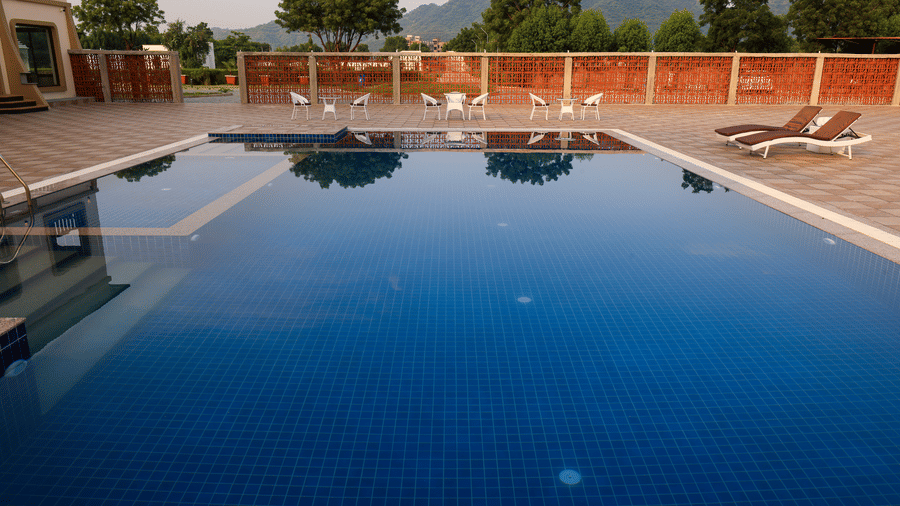 A calm, blue swimming pool reflecting the sky, surrounded by a stone deck with lounge chairs and a red privacy wall - Tathastu Chavri Resort, Jawai