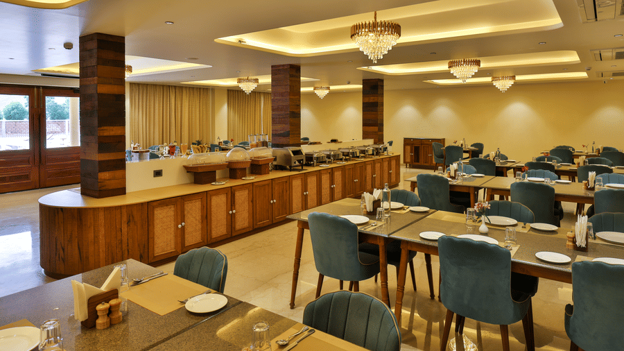 An expansive indoor restaurant with multiple wooden dining tables, blue chairs, and warm recessed lighting - Tathastu Chavri Resort, Jawai