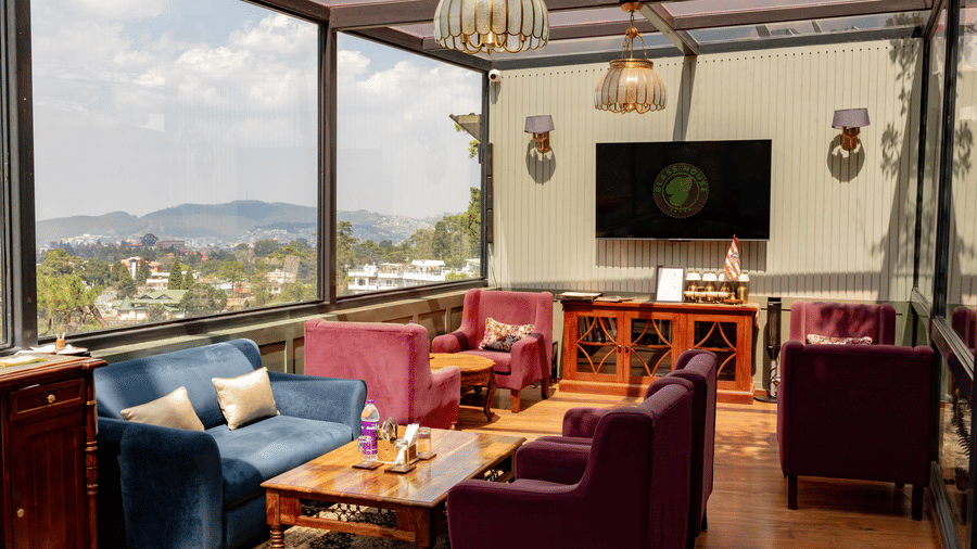 A dining space at The Heritage Club, Tripura Castle, Shillong, with lamps, tables, cushions, and a wall-mounted television showcasing, Glass House. 