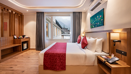 A hotel bedroom at Lords Inn Katra featuring a king size bed, a side table with a telephone, and a glass window showing adjacent structures.