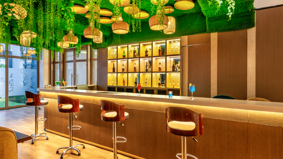 Wide view of the bar at Lords Inn Sangam, Bhopal with a curved wooden counter, red stools, bottle display shelves, and green ceiling decor.