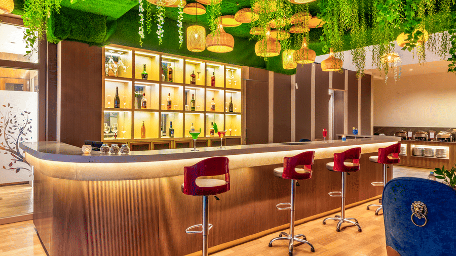 Elegant curved bar at Lords Inn Sangam, Bhopal featuring red and blue stools, illuminated liquor shelves, and a decorative green ceiling.