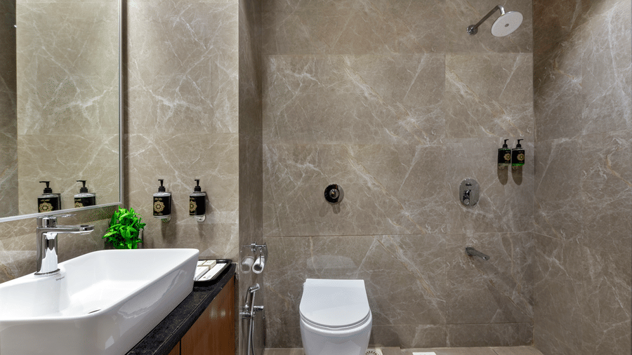 A modern bathroom in Lords Somnath, with sleek marble walls, featuring a walk-in shower, wall-mounted toilet, large mirror, and counter top basin. 