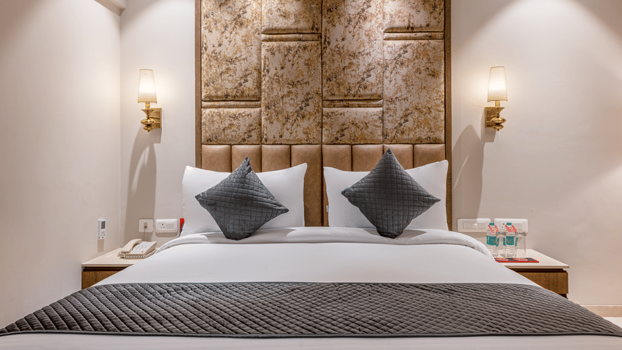 A close up of a double bed in the Deluxe Room with grey and white linens, set against a decorative carved wooden headboard – Skkylight Lords Inn, Nanded