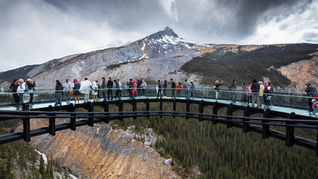 Tourists standing on a curved glass-floored skywalk overlooking a forested canyon, with a snow-capped mountain peak rising in the background under a cloudy sky.