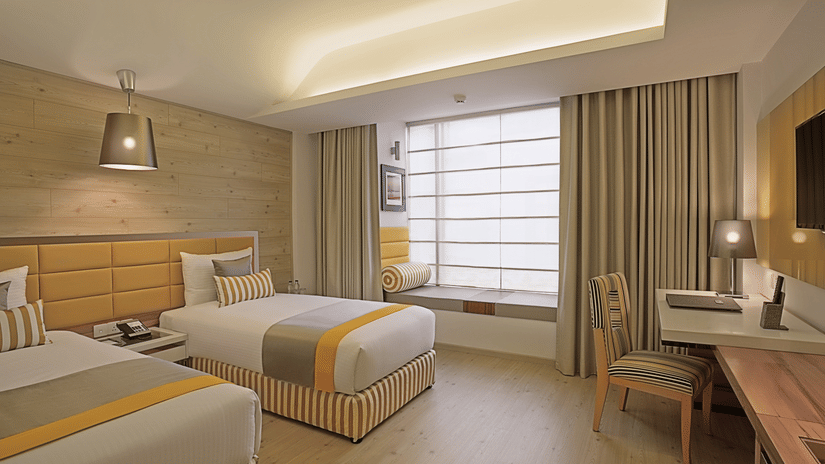 An Deluxe Twin Room at Barsana Boutique Hotel, Kolkata, featuring twin beds draped in matching linens, a large window, and a workspace with a chair.