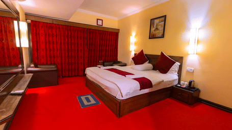 A bedroom with a king size bed, 2 arm chairs, a large mirror, a carpet, a bedside table, wall lamps, and wall art at Hotel Seagull Digha.