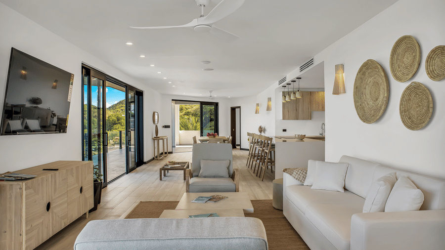 A living room with a sofa, a lounge chair, a side sofa, a mounted television, a cabinet, a dining area,a kitchen area and two glass sliding doors at Tamarind Hills Resort and Villas, Antigua.