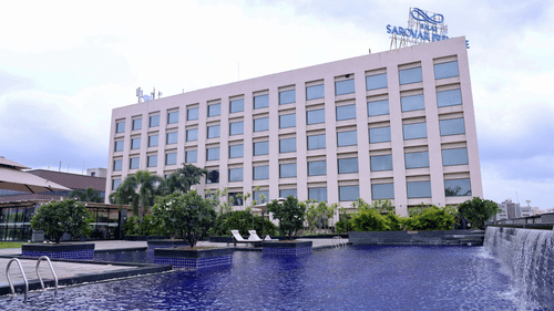 A hotel building with a pool in the foreground, featuring water features and lounge chairs.