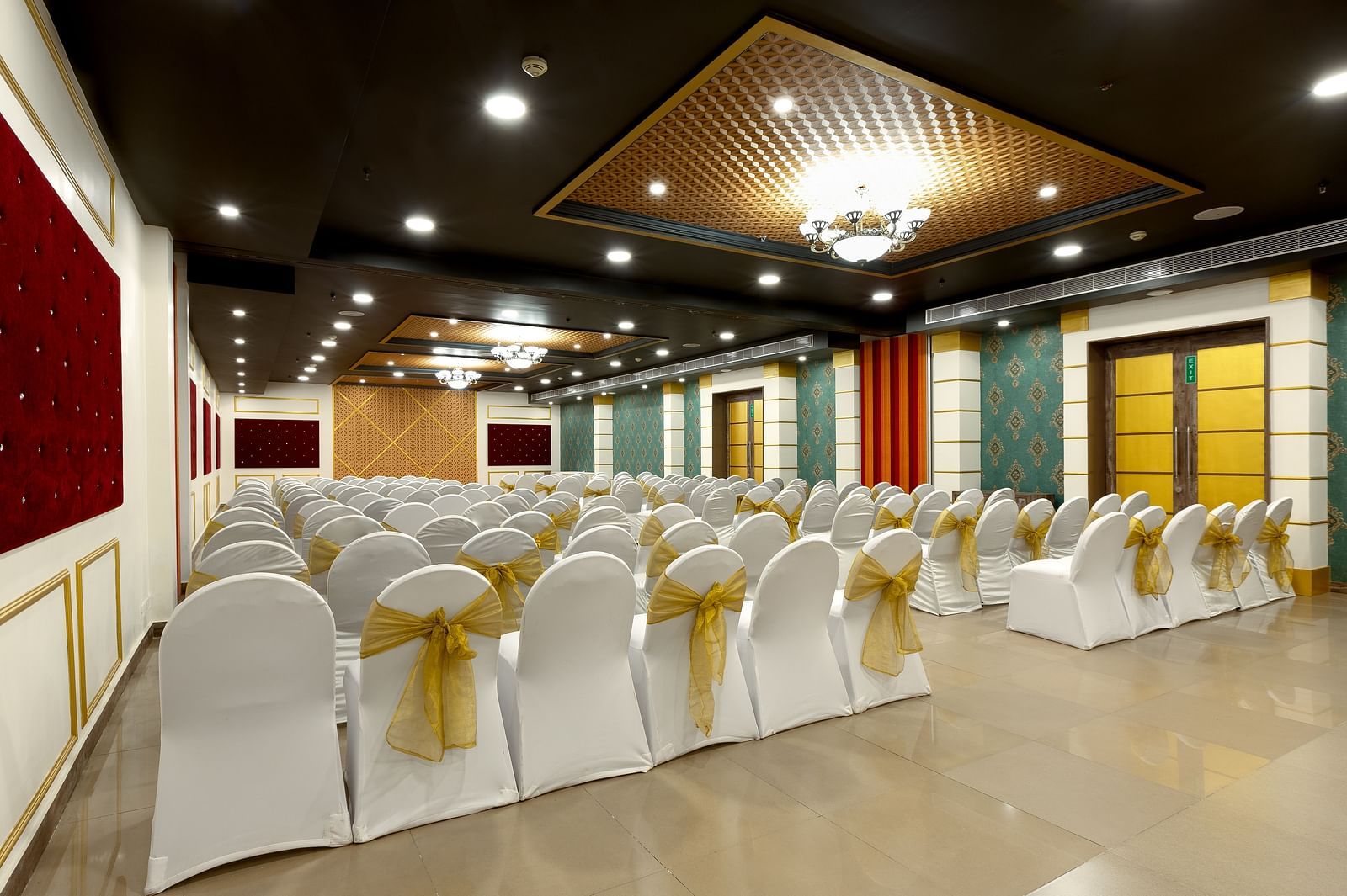 A spacious banquet hall with chairs covered in white cloths and gold  covers. The hall has a modern design with dark wood accents and a crystal chandelier.