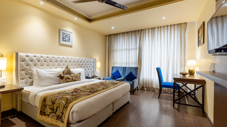 A bedroom with a king size bed, side tables, work desk, chair, lamps, curtains, and wall-mounted TV at Indraprastha Resort & Spa, McLeod Ganj.