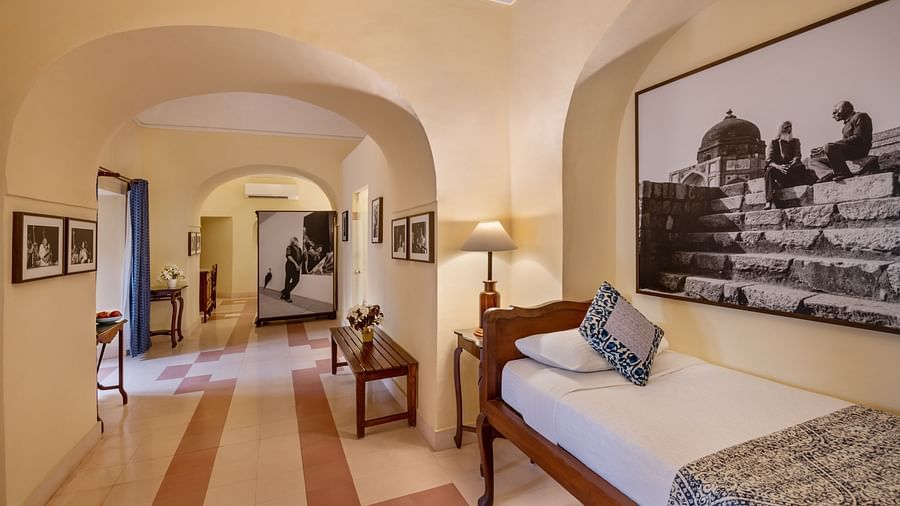 A room with a single bed, a framed artwork, and an arched doorway leading to another room in Tijara Fort-Palace - 19th Century, Alwar.